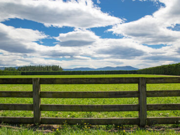 Weite Landschaft mit grünem Feld, Holzzaun und bewölktem Himmel über Bergen im Hintergrund.