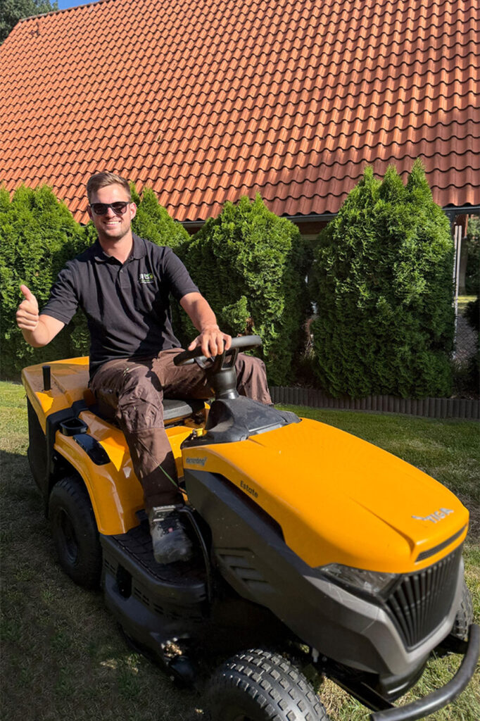 Ein Mann fährt einen Rasenmäher-Traktor auf einem gepflegten Grundstück mit Bäumen im Hintergrund.
